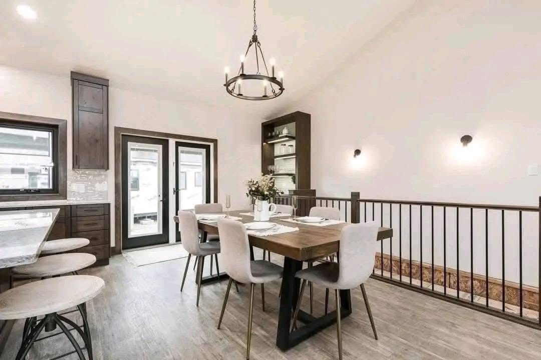 Spacious modern living area with open kitchen, dark wood cabinets, island, and hanging lights. Cozy seating with gray armchair, black sofa, white rug.