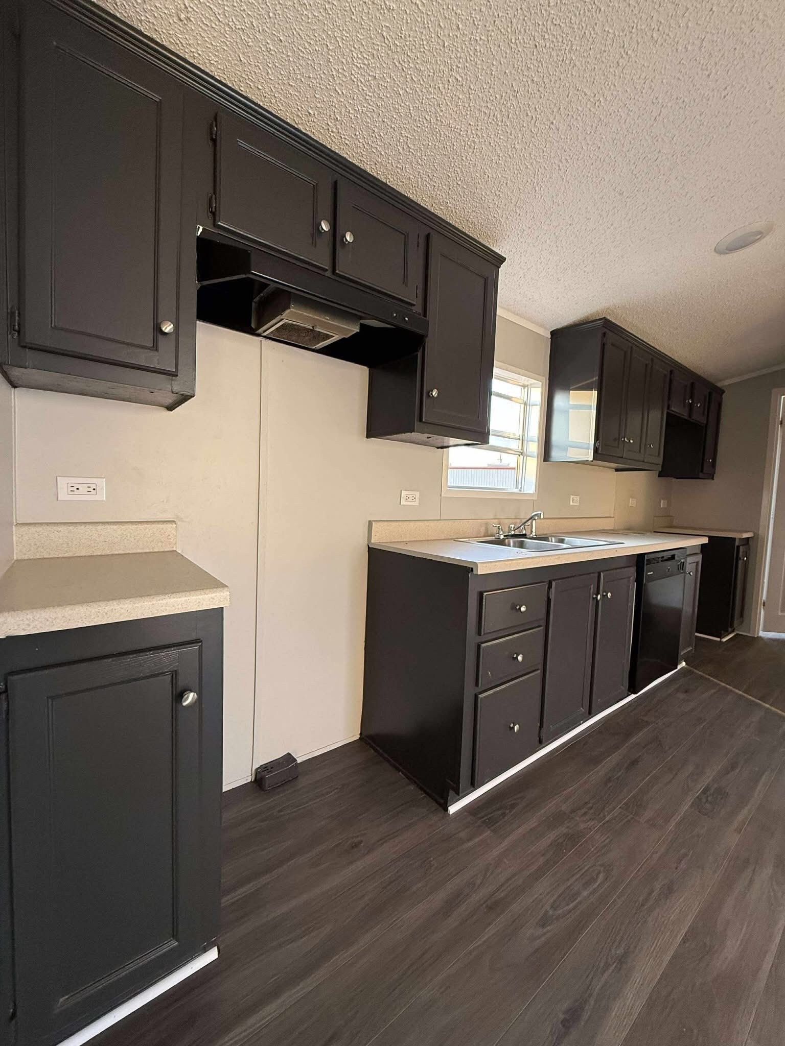 Modern kitchen with dark cabinets, stainless steel sink, and beige countertops. Wood flooring contrasts with light walls, creating a sleek, clean look.