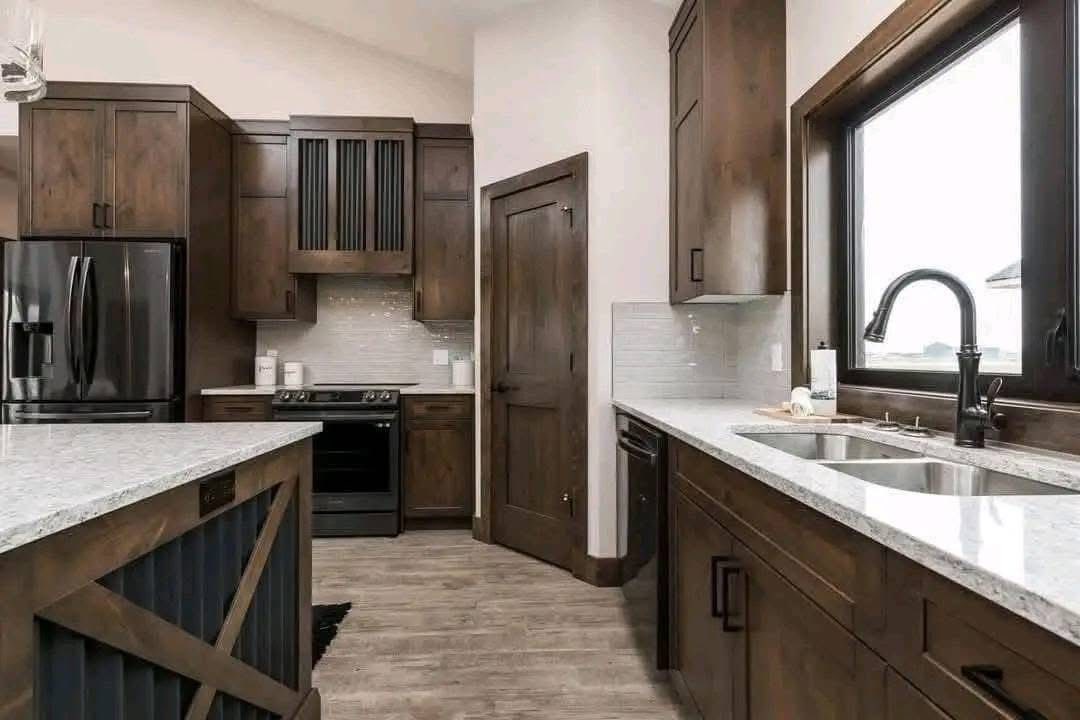 Modern kitchen with dark wood cabinets, a black fridge, and stainless steel appliances. Light countertops and windows create a bright, inviting atmosphere.