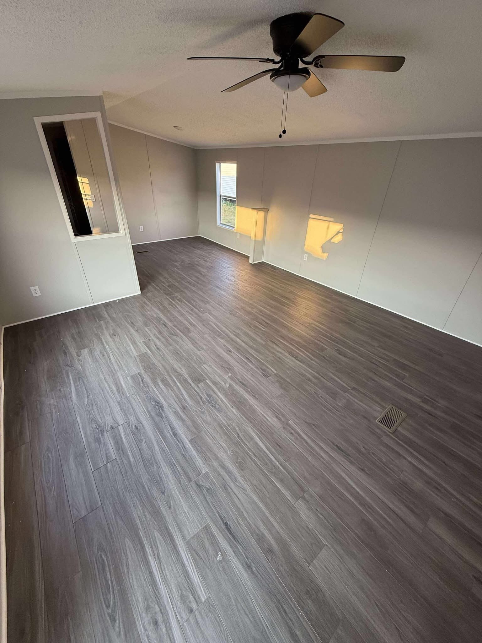 Empty room with gray wood flooring, light gray walls, and a ceiling fan. Two windows allow soft natural light, creating a calm atmosphere.
