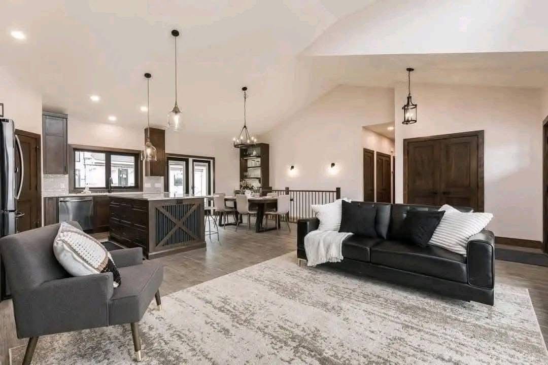 Spacious modern living area with open kitchen, dark wood cabinets, island, and hanging lights. Cozy seating with gray armchair, black sofa, white rug.
