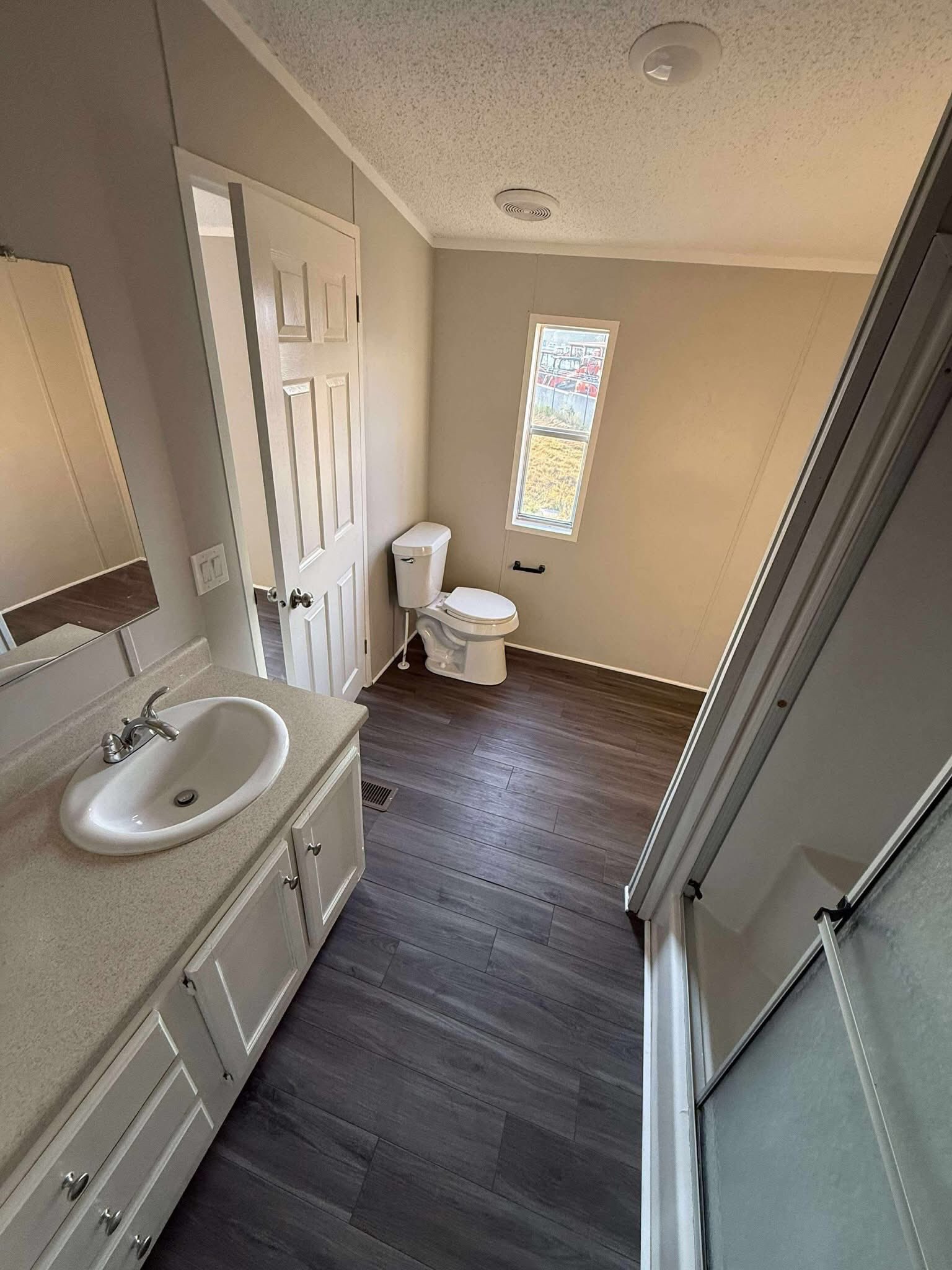 Compact bathroom with light beige walls and dark wood floor. Features include a sink with cabinet, toilet, small window, and shower, creating a clean and minimal look.