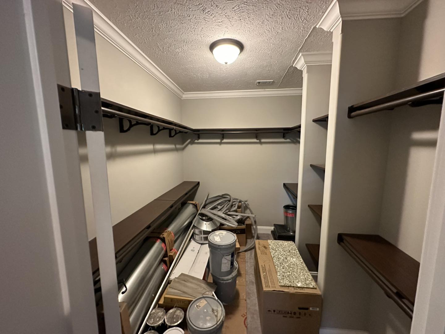A walk-in closet under renovation with bare shelves and a beige wall. The floor is cluttered with construction materials, boxes, and paint cans, creating a work-in-progress feel.
