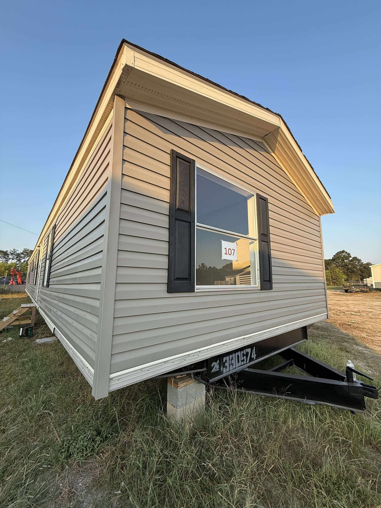 Beige mobile home with black shutters on a sunny day, labeled "107." It stands on a grassy lot with a trailer hitch visible in the foreground.