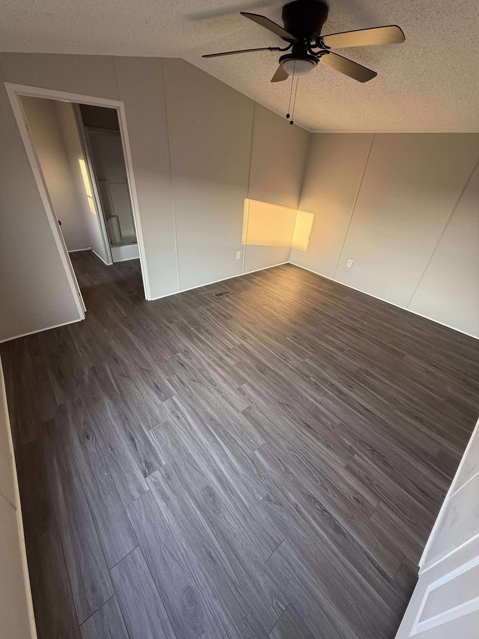 Empty room with dark wood flooring, beige walls, and a ceiling fan. Sunlight streams through a window, creating a warm and inviting ambiance. A doorway opens to a small bathroom.