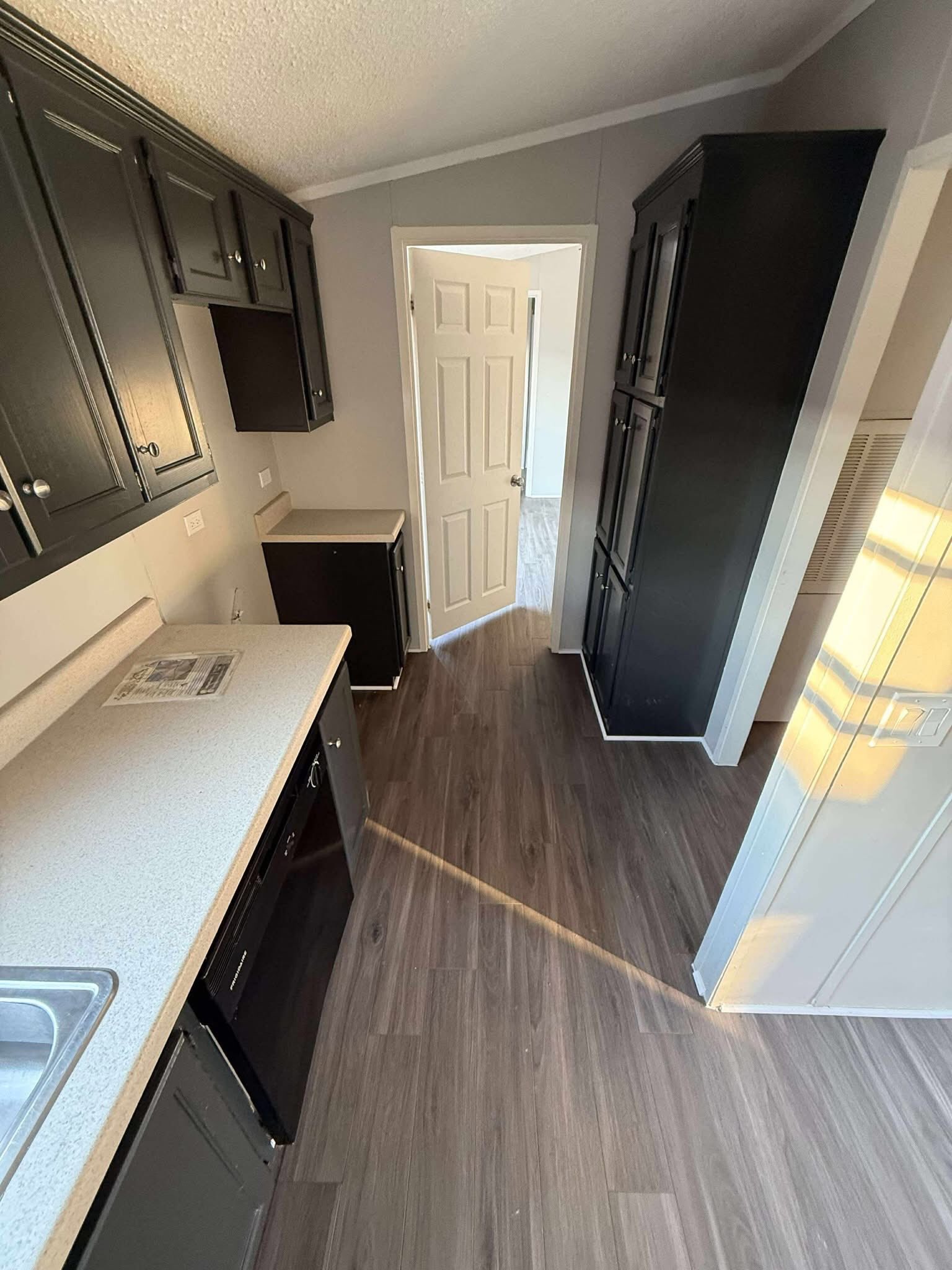 Narrow kitchen with dark cabinets and light countertops. Sunlight streams through a window, creating a cozy, inviting atmosphere.