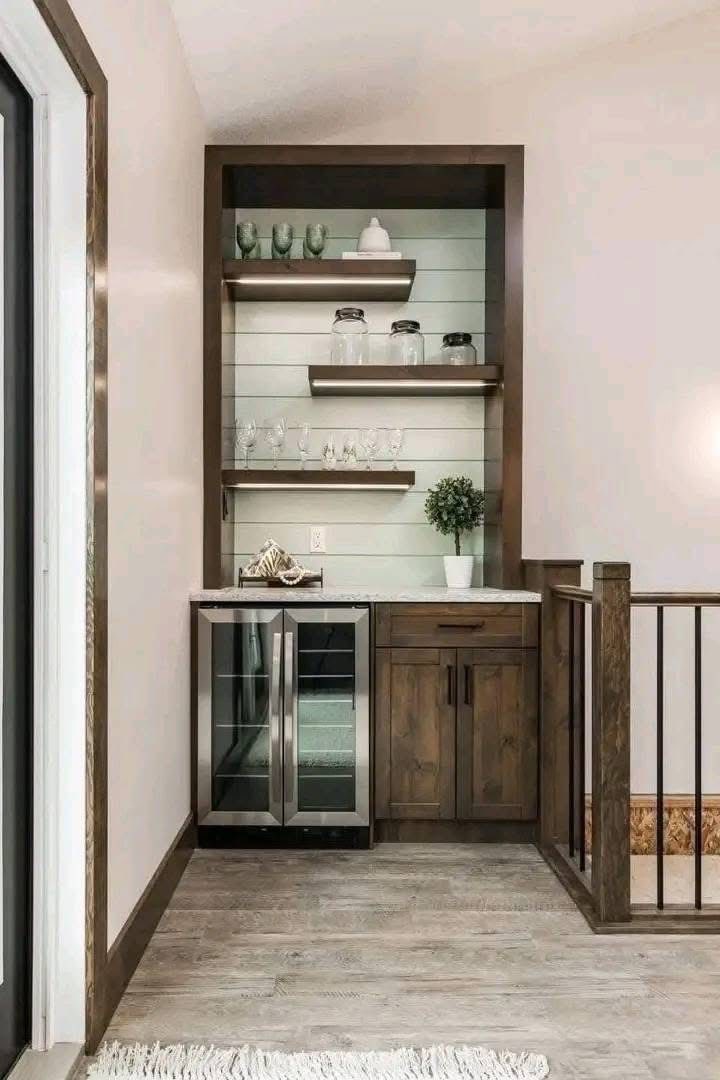 Compact home bar with rustic wooden shelves holding green mugs, glass jars, and glasses. Below, a mini fridge and cabinet with a potted plant create a cozy, modern vibe.