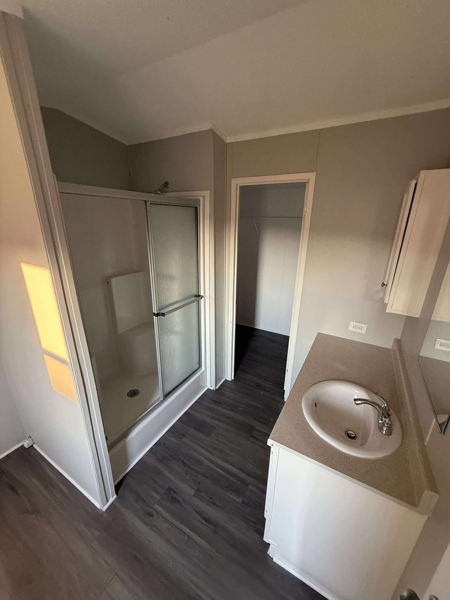 A small bathroom with gray walls features a glass-enclosed shower, wood flooring, a beige countertop with a sink, and a cabinet. The lighting is warm and soft.