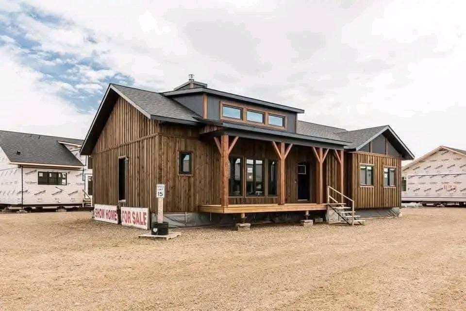 Wooden show home for sale on a dirt lot, featuring large windows and a small porch. Other modular homes visible in the background under a cloudy sky.