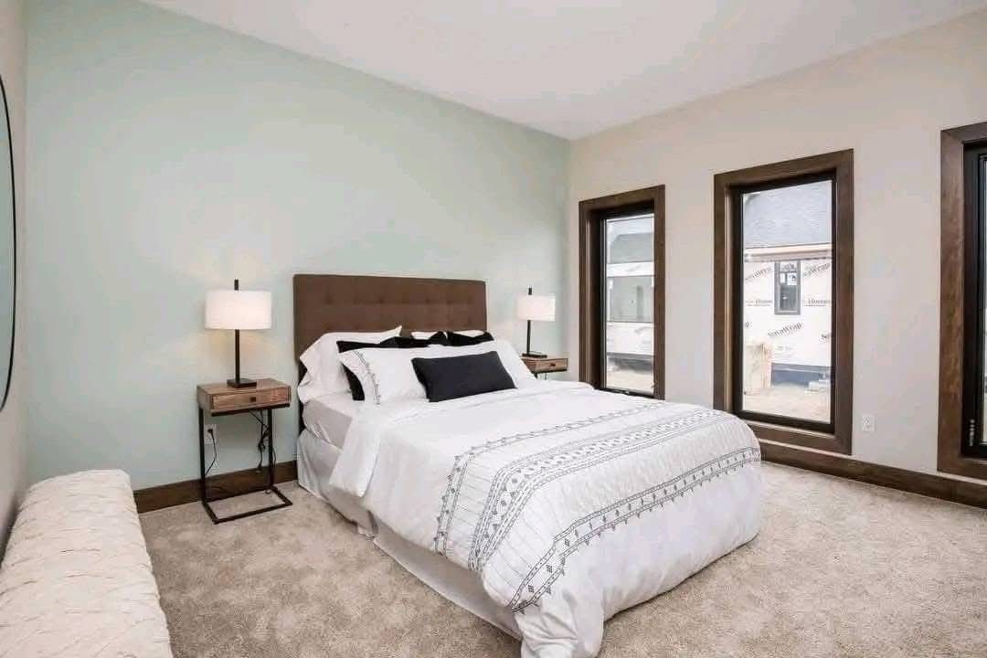Modern bedroom with a soft color palette features a neatly made bed with white and black accents between two wooden nightstands with lamps. Three large windows allow natural light to brighten the space, creating a calm and inviting atmosphere.