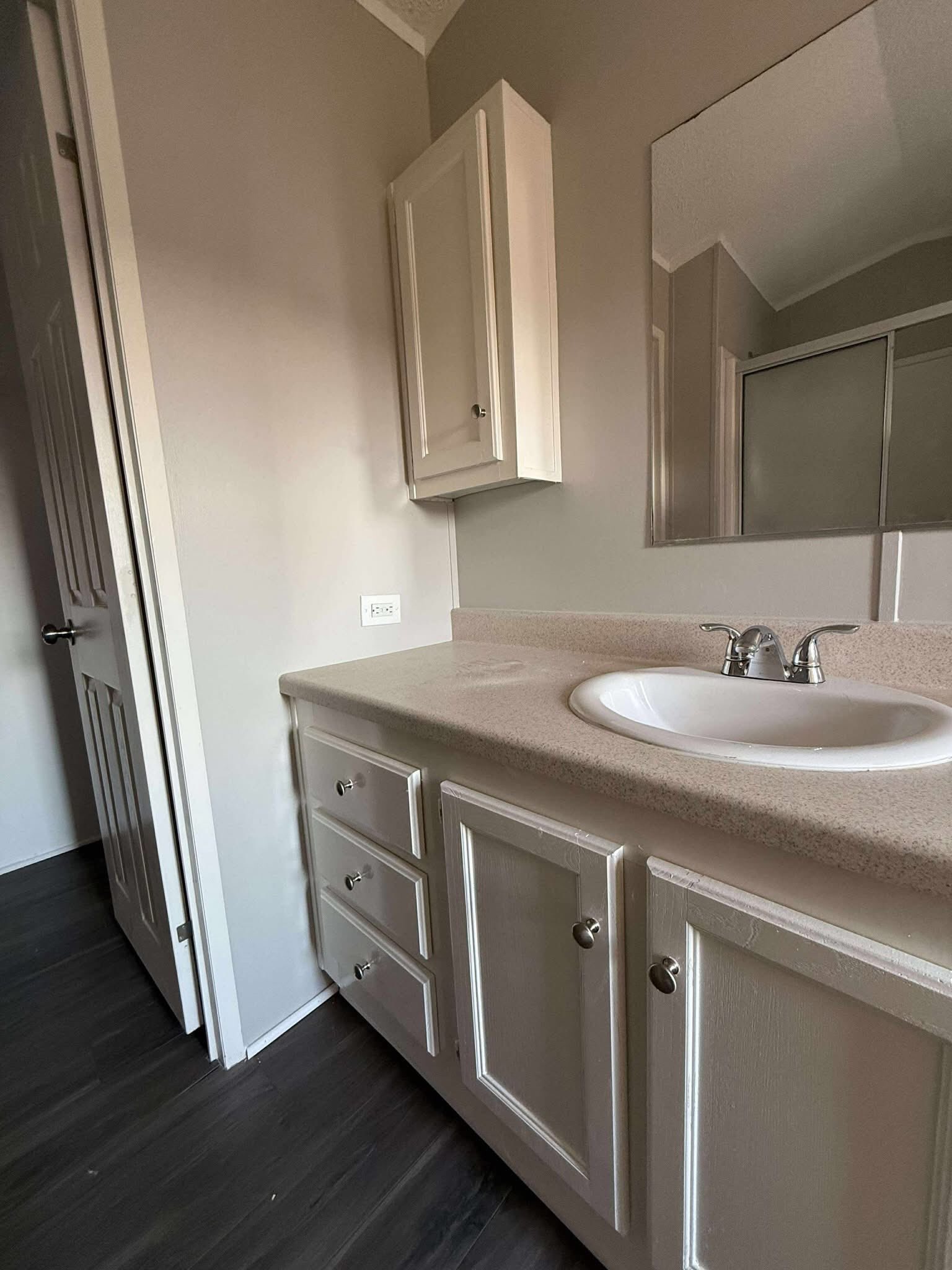 Neutral-toned bathroom with dark flooring, white vanity, cabinets, and silver faucet. Large mirror reflects the space, adding depth and light.