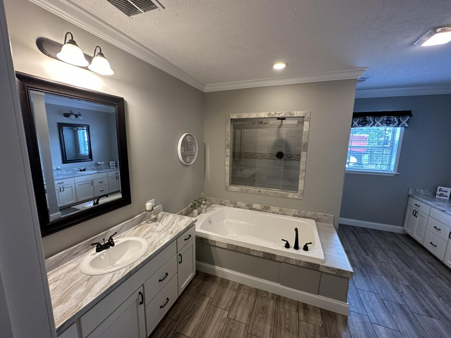 Modern bathroom with gray walls, featuring a marble countertop and sink, large mirror, bathtub with faucet, and wood-look tile flooring.