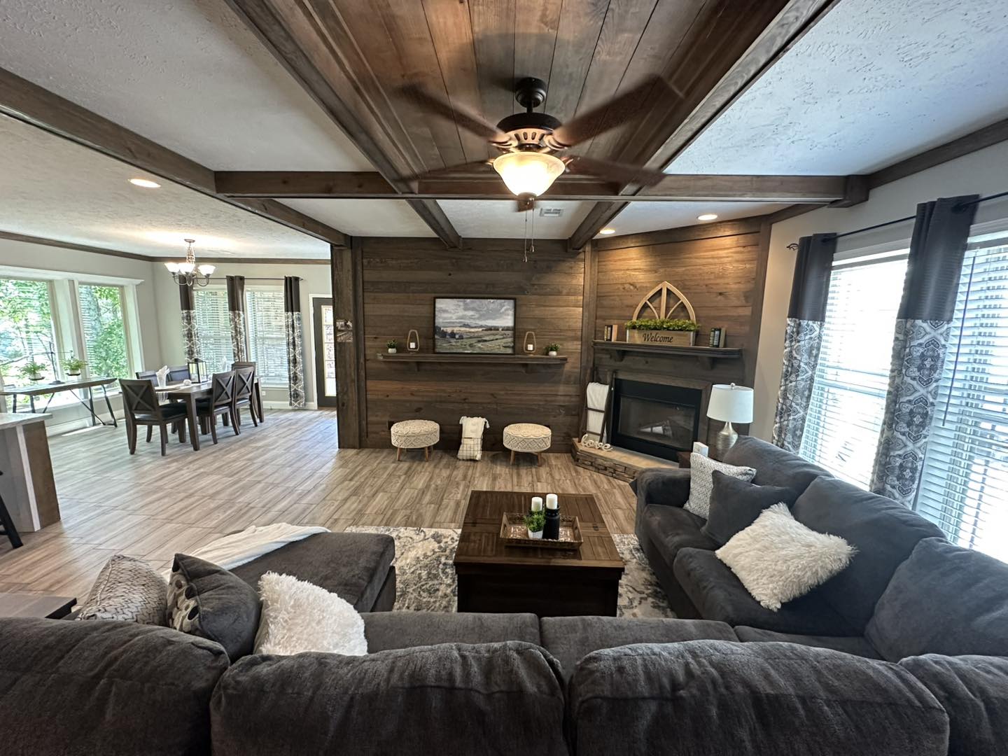 Spacious living room with dark gray couches, wooden beams, and a ceiling fan. Features a cozy fireplace, large windows, and adjoining dining area.