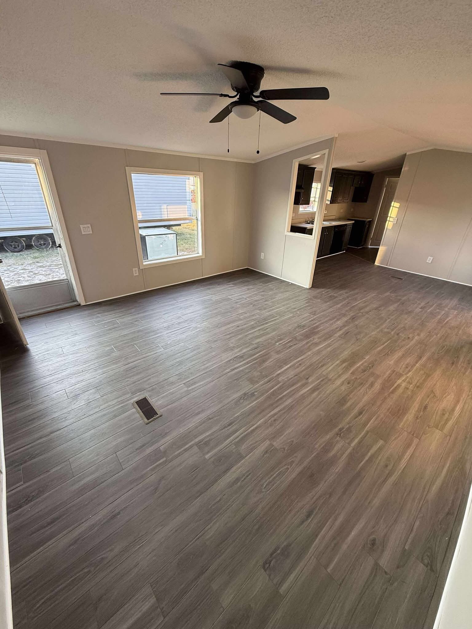 Spacious empty room with dark wood flooring and beige walls, featuring a ceiling fan and large windows. The room opens to a modern kitchen.