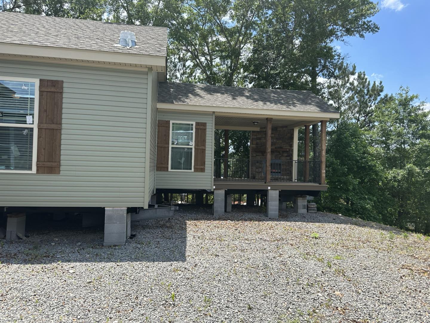 A beige modular home elevated on cinder blocks with a stone-accented porch. Surrounded by trees under a clear blue sky, creating a peaceful outdoor setting.
