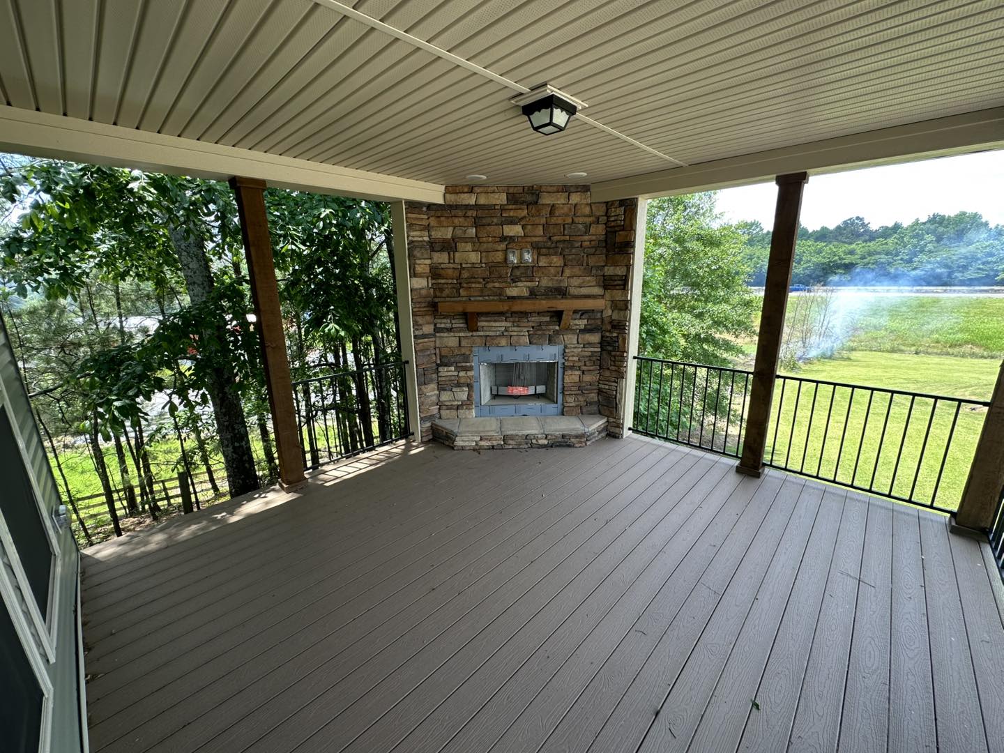 Covered porch with stone fireplace, wooden beams, and black railing. Overlooks lush green trees and a serene pond, conveying a cozy, peaceful ambiance.