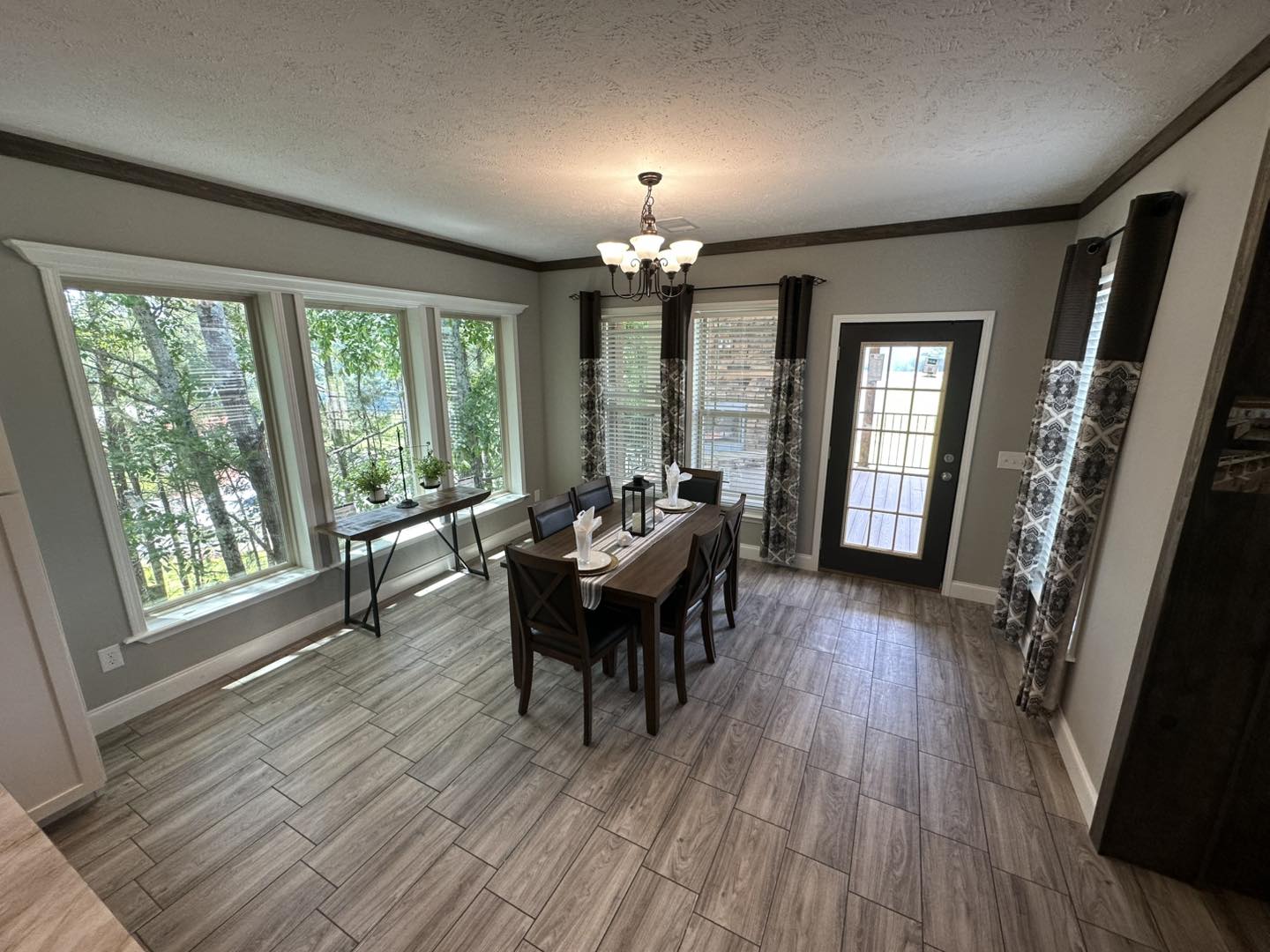 Dining room with large windows and a glass door allowing natural light. A wooden table set for four, modern chandelier, and tiled floor create a welcoming ambiance.