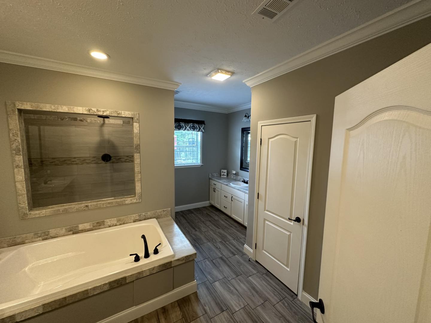 A modern bathroom with gray walls and wood-like tile flooring. Features a large bathtub with black fixtures, a glass shower, and a white vanity under a window.