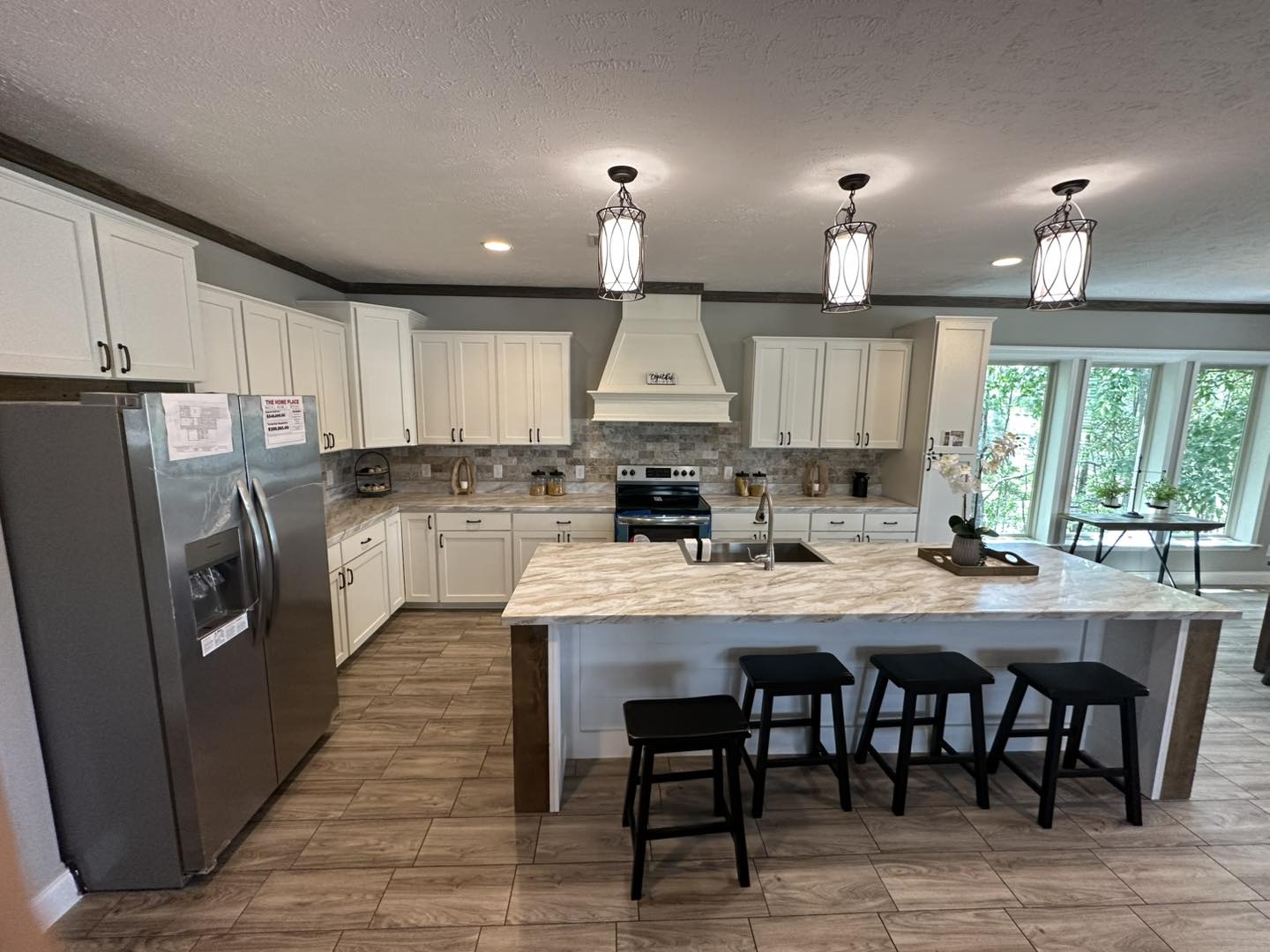 Modern kitchen with white cabinets, stainless steel appliances, and a marble island. Three pendant lights hang above four black stools, creating a welcoming atmosphere. Large windows on the right let in natural light.