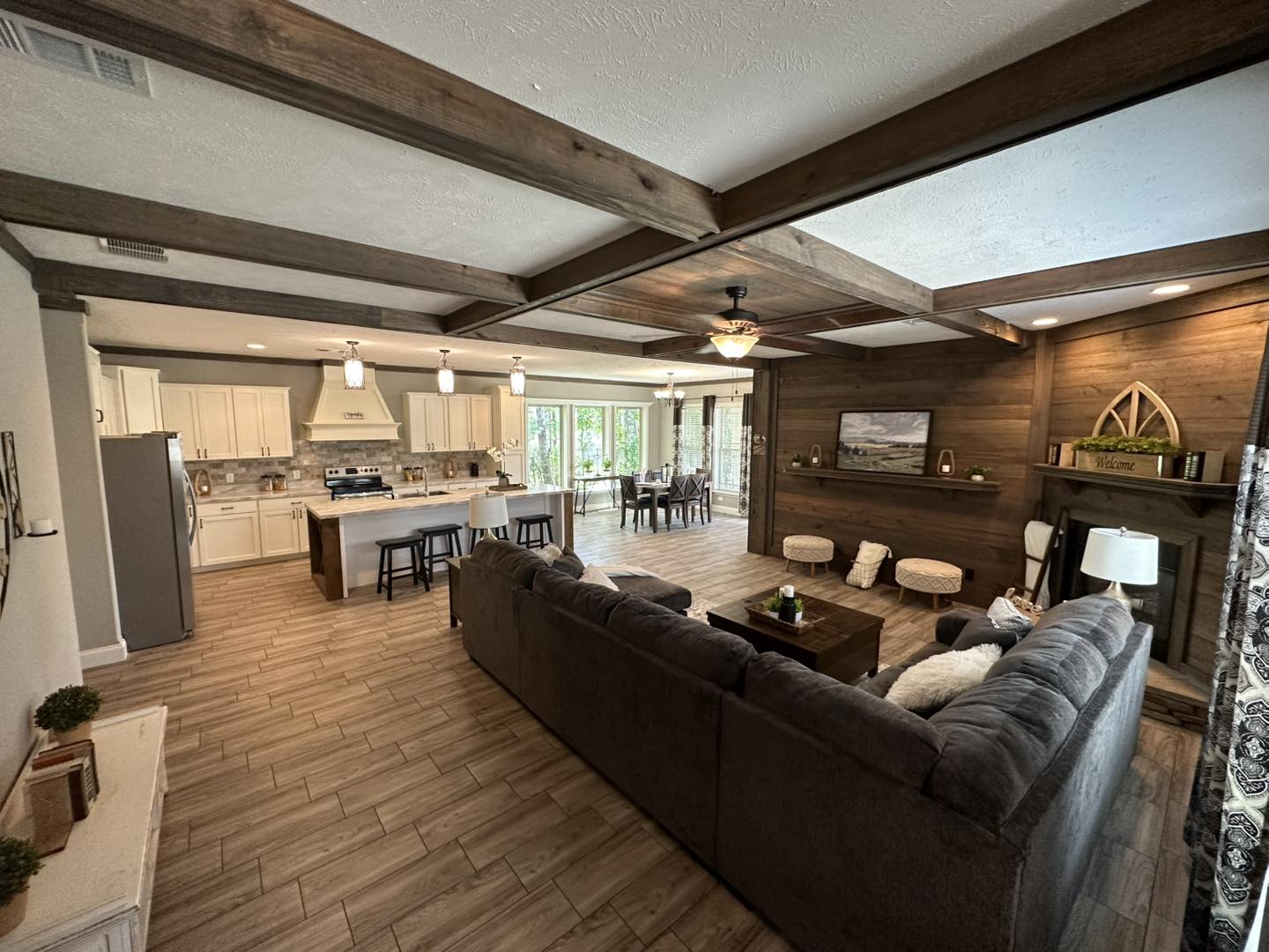 Cozy open-plan living area with rustic wooden beams, a kitchen with pendant lights, dining table by large windows, and a gray sectional sofa by a wooden accent wall.