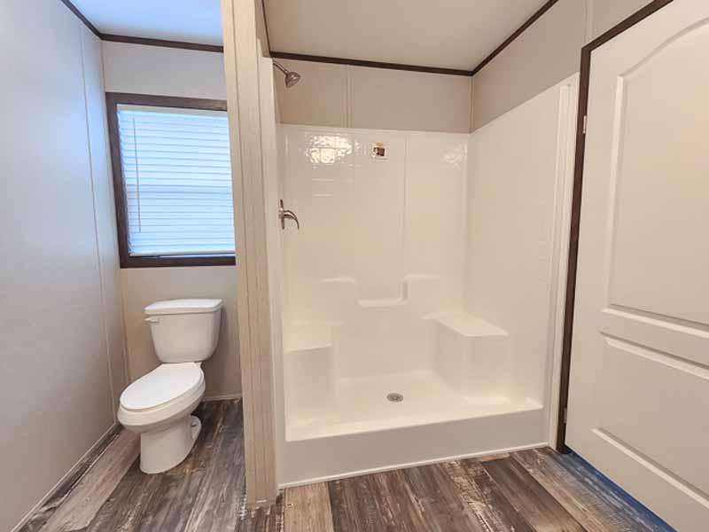 A clean bathroom with wood-patterned floor, featuring a white toilet beside a window with blinds, and a spacious white shower with a glass door.