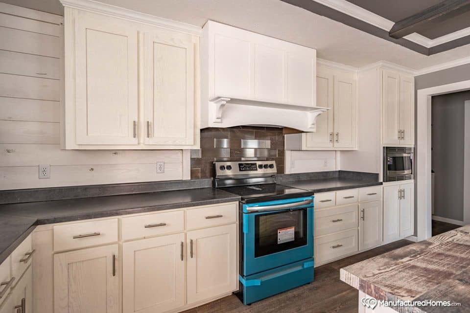 Modern kitchen with cream cabinets and dark stone countertops. A striking stainless steel stove with a blue protective covering and a sleek microwave are featured. The warm brown backsplash and rustic wooden table create a cozy, inviting atmosphere.