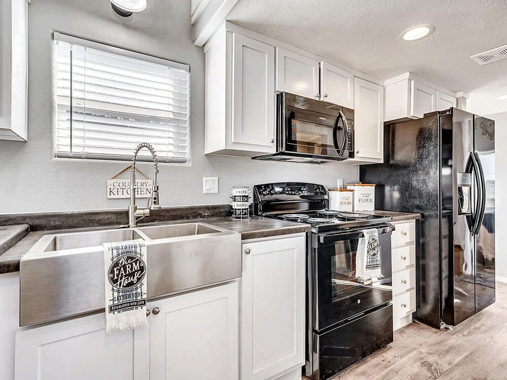 Modern farmhouse kitchen with white cabinets, black appliances, and a large stainless steel sink. Sunlight streams through a window, creating a warm, inviting atmosphere. A "Farm House" towel hangs on the sink.
