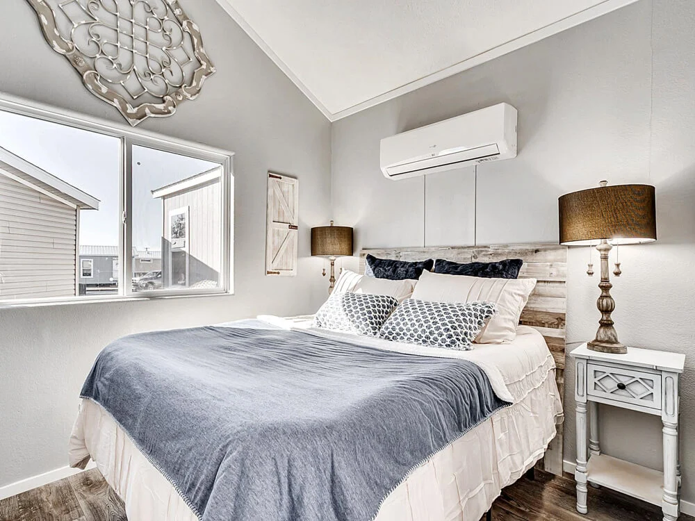Cozy bedroom with a rustic wood headboard, white and blue bedding, dual bedside lamps, and an air conditioner above. Large window with natural light.