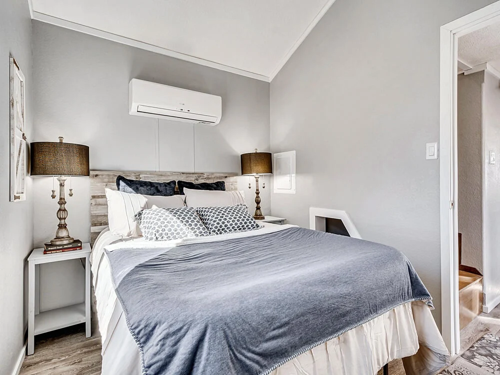 Cozy bedroom with gray walls features a neatly made bed with a blue blanket and patterned pillows, flanked by two side tables with black lamps. A wall-mounted air conditioner is above the headboard, adding modern comfort. The space feels calm and inviting.