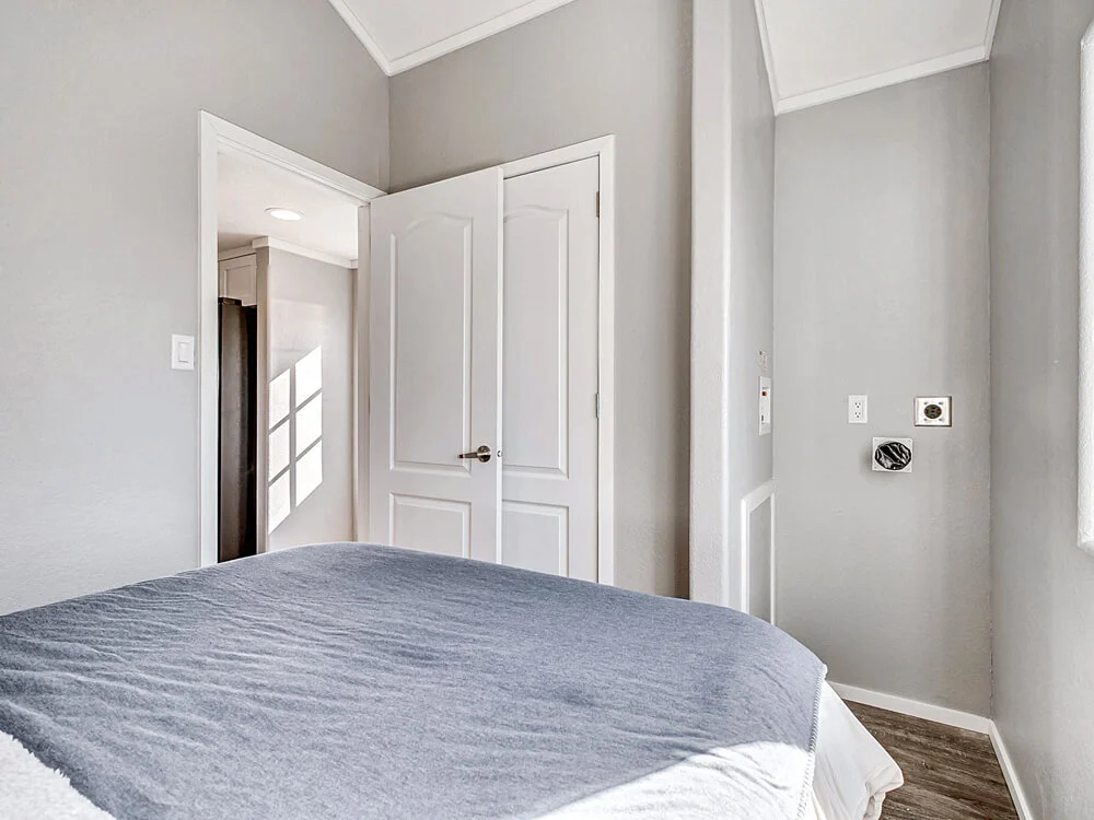 Cozy bedroom with gray walls and bedspread. A door is slightly open, revealing light from an adjacent room. Sunlight casts window shadows.