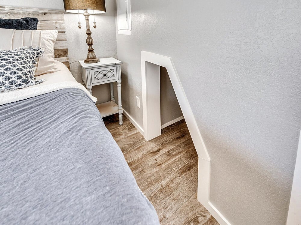 Cozy bedroom with wooden floors, a bed with gray bedding, a patterned cushion, a small white nightstand with a lamp, and a tiny doorway on the wall.