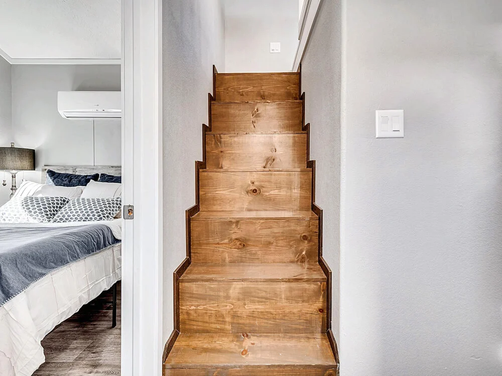 Wooden stairs with a modern, narrow design lead upwards in a small hallway. On the left, a glimpse of a cozy bedroom with a made bed. The tone is warm and inviting.