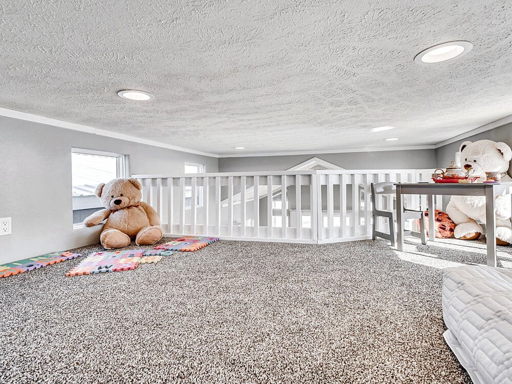 Cozy children’s play loft with carpet, large teddy bears, colorful play mats, and a small table. Bright and inviting with recessed lighting.