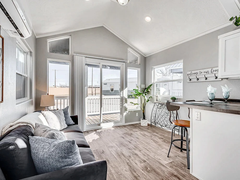 Bright, modern living room with vaulted ceilings, gray walls, and wood flooring. Features a gray sofa, bar area, and large windows with patio view. Cozy and inviting.