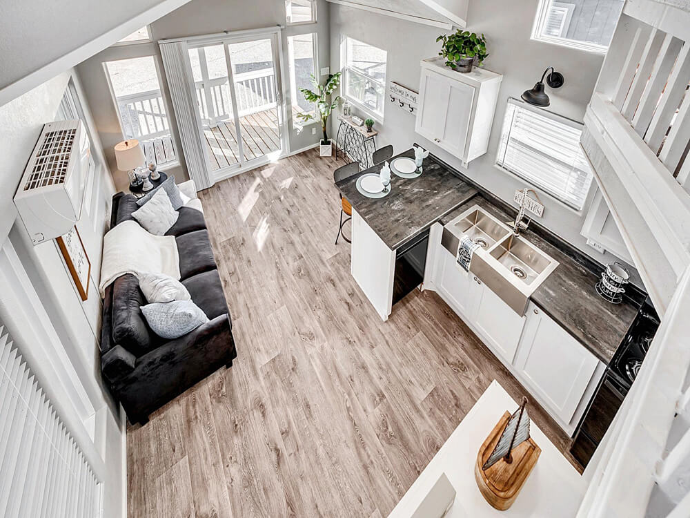 Aerial view of a modern, airy living room and kitchen. Features include a gray sofa, wood floors, white cabinets, and ample natural light from large windows.