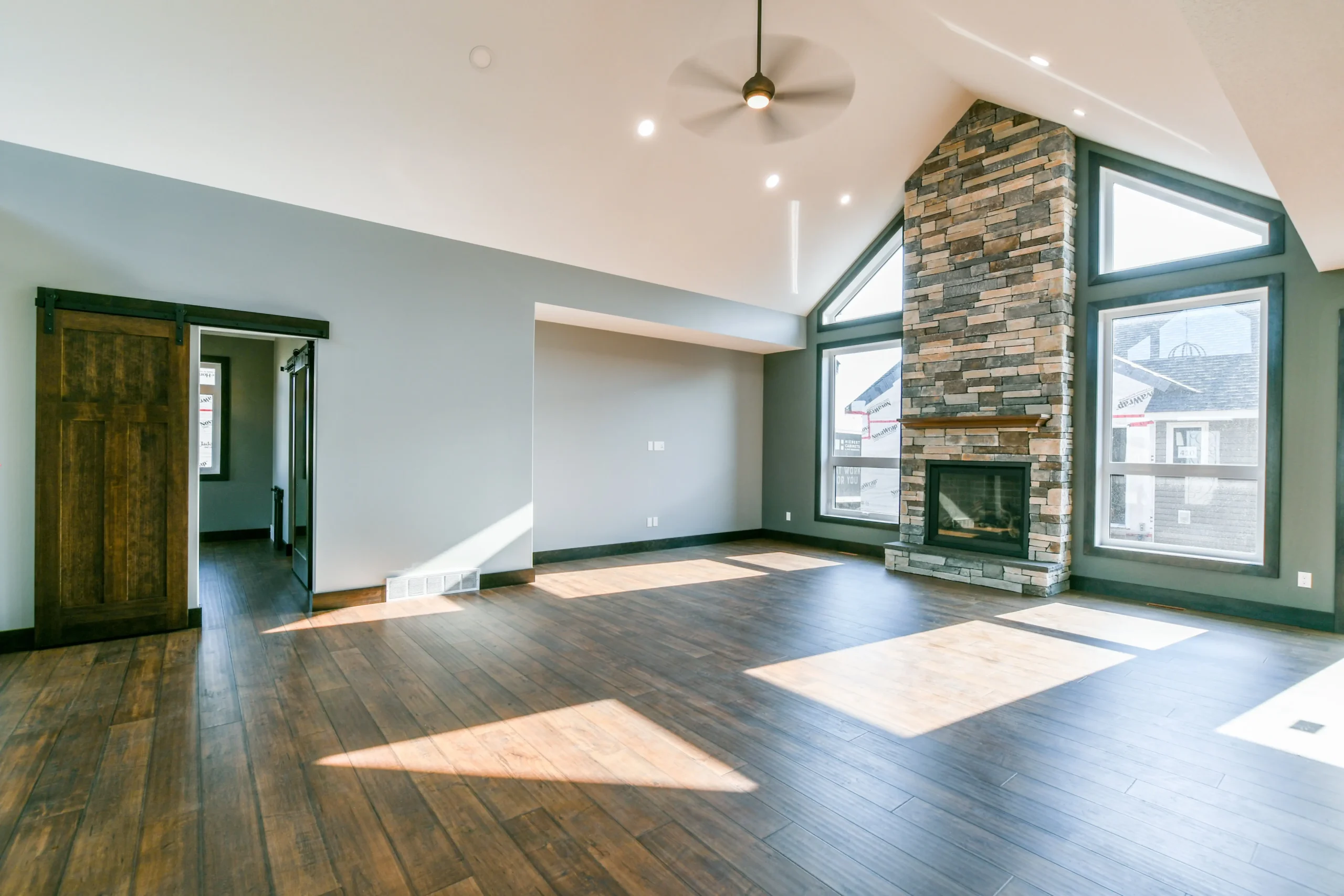 Spacious, well-lit living room with dark hardwood floors, stone fireplace, and large windows. A sliding barn door adds a rustic touch.