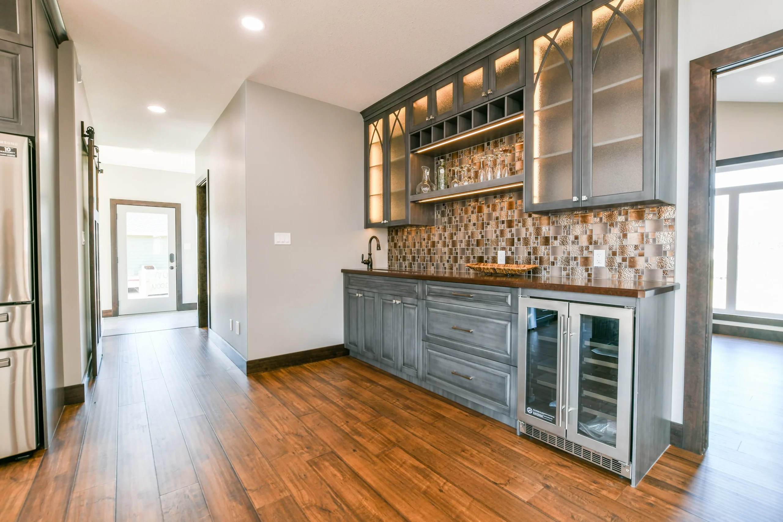 Stylish kitchen with wood flooring, grey cabinets, and a mosaic tile backsplash. It features open shelves with glassware and a wine fridge. Bright and modern.