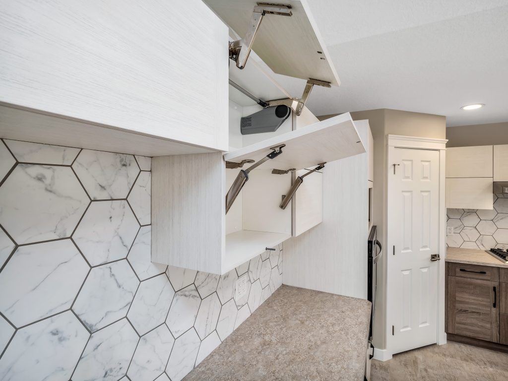 Modern kitchen with white hexagonal marble backsplash, open white upper cabinets with hydraulic lifts, beige countertop, and closed pantry door. Bright and sleek.
