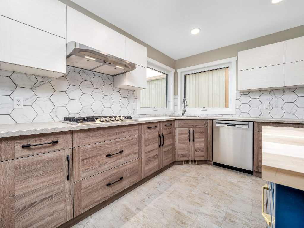 Modern kitchen with light wood cabinets, black handles, hexagonal white tile backsplash, gas stove, and stainless steel dishwasher. Bright and clean ambiance.
