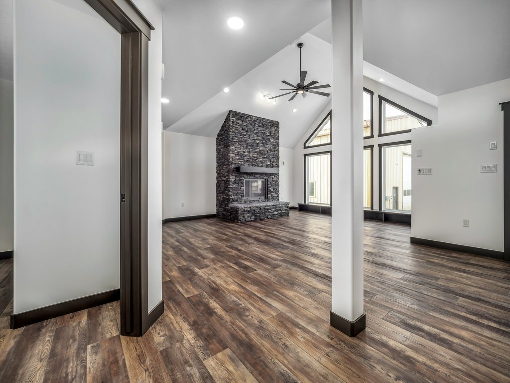 Spacious modern living room with vaulted ceiling, large windows, and a central stone fireplace. Warm wood floors and neutral walls create an inviting feel.
