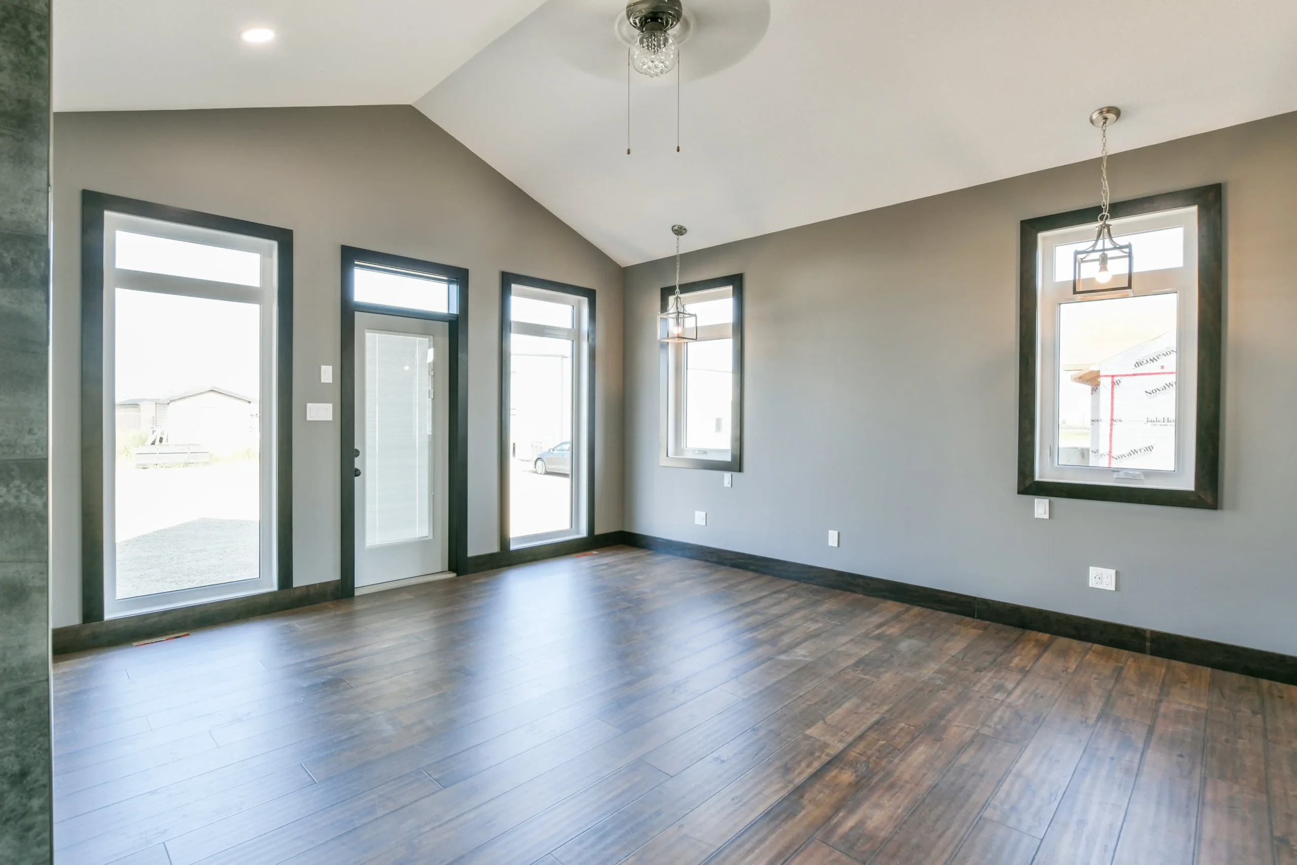 Spacious, empty living room with wood flooring, gray walls, and high ceiling. Large windows and glass door allow natural light. Modern pendant lights.