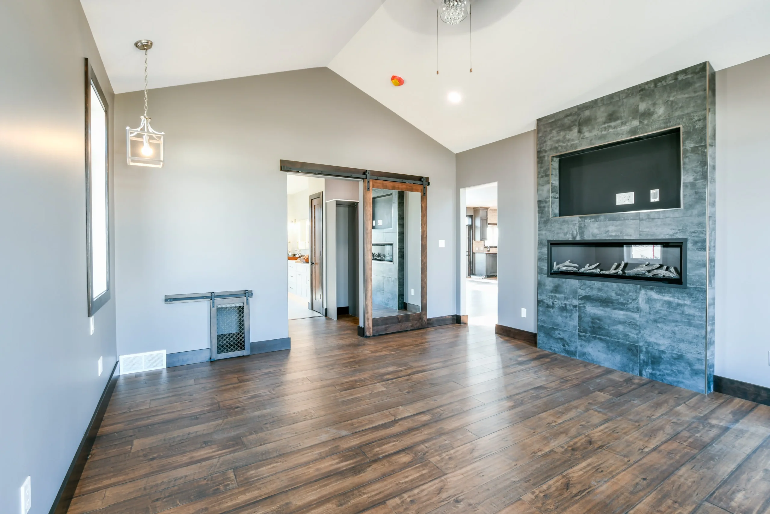 Spacious modern living room with dark wooden floors, gray walls, and a minimalist fireplace. A sliding barn door opens to the kitchen, enhancing the open feel.