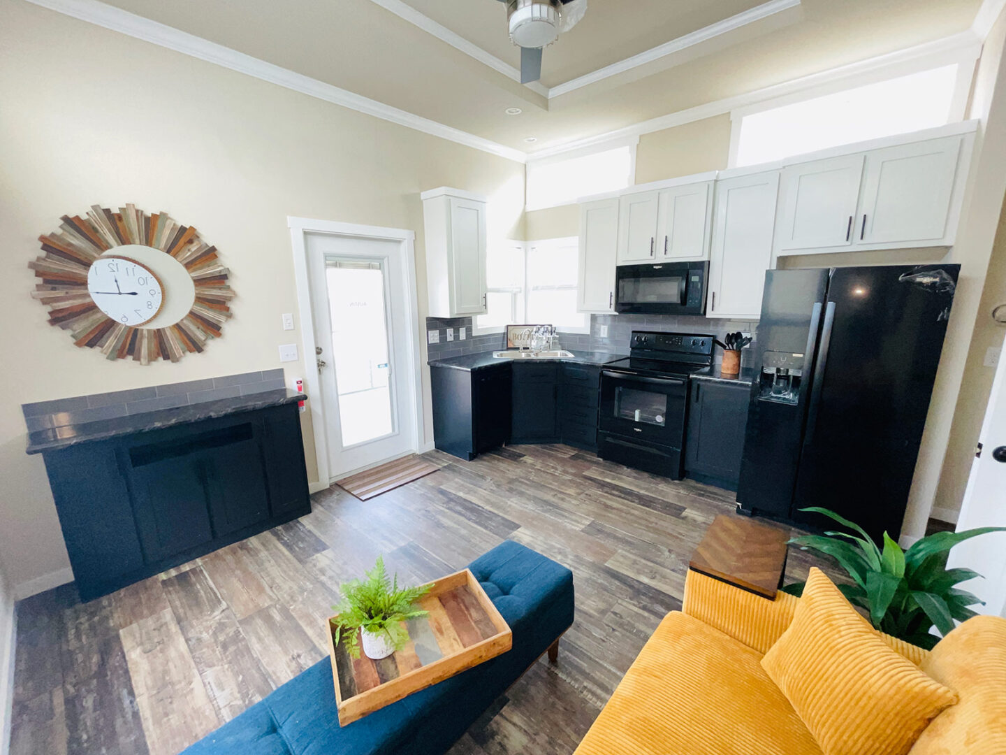 Modern kitchen and living area with wood floors, dark cabinets, and white upper cabinets. A yellow couch and a blue ottoman add vibrant color.