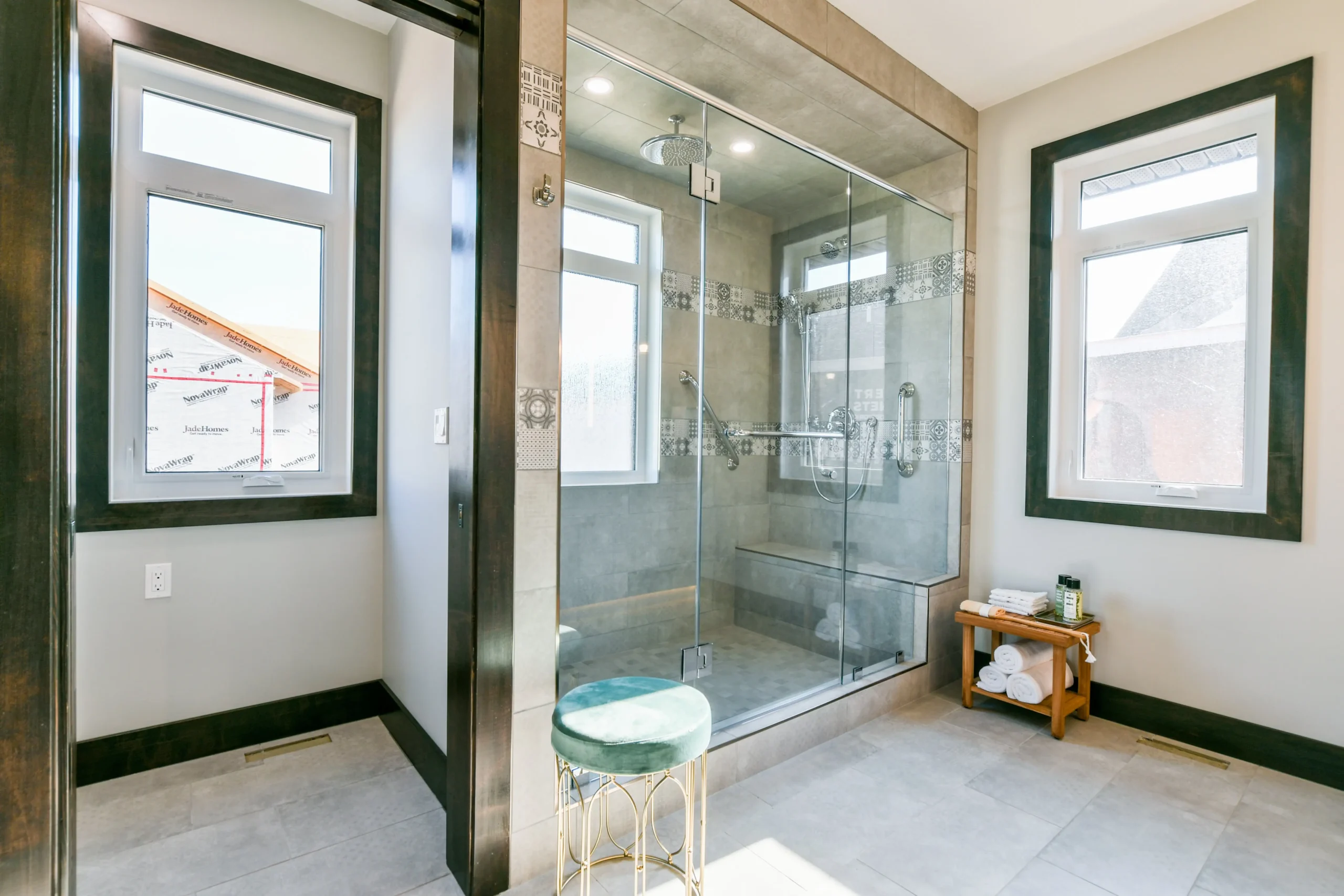 Modern bathroom with a large glass-enclosed shower featuring gray tiling and a rain shower head. Light floods through two windows, illuminating a small wooden bench with towels and toiletries. A teal cushioned stool adds a touch of elegance.