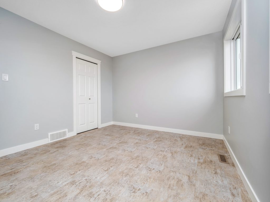 An empty room with light gray walls, a wooden floor, and a white door. A window on the right lets in natural light, creating a fresh, clean atmosphere.