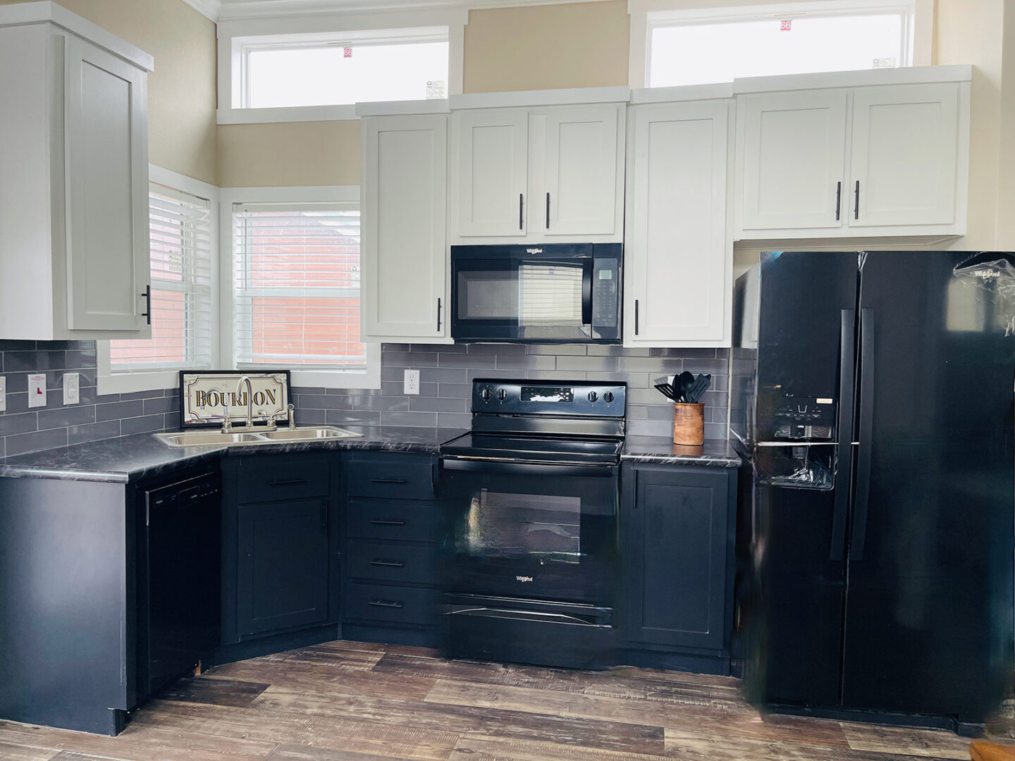 Modern kitchen with black and white cabinets, black appliances, and gray tile backsplash. Natural light from small windows gives a clean, sleek feel.