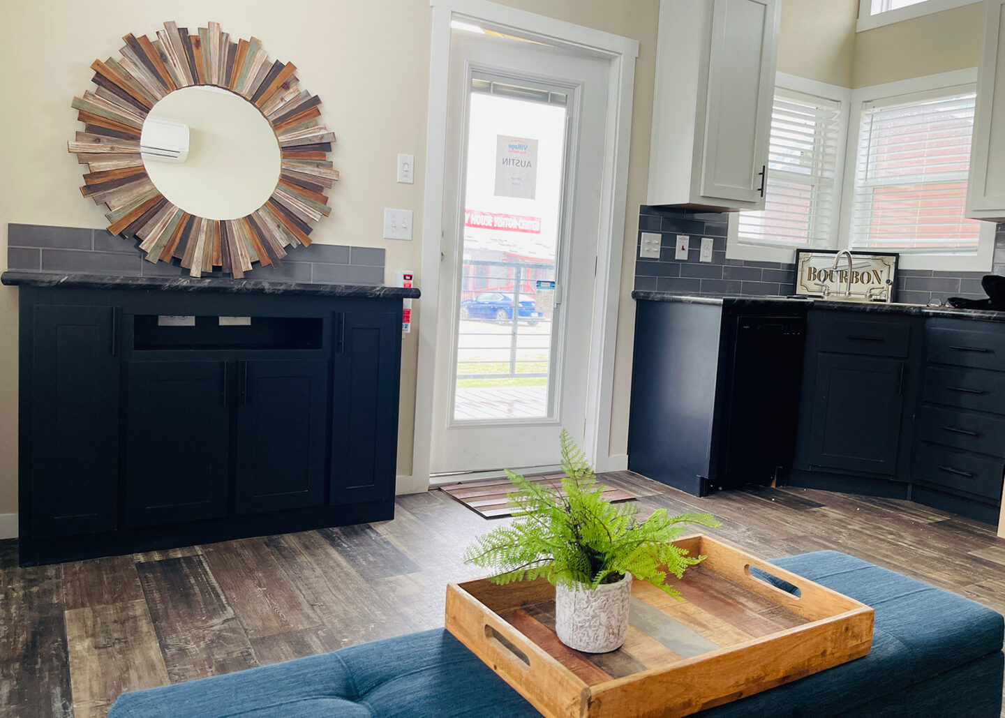 Cozy living space with a rustic mirror above a black cabinet. A wooden tray with a fern sits on a blue sofa. Bright light filters through a glass door.