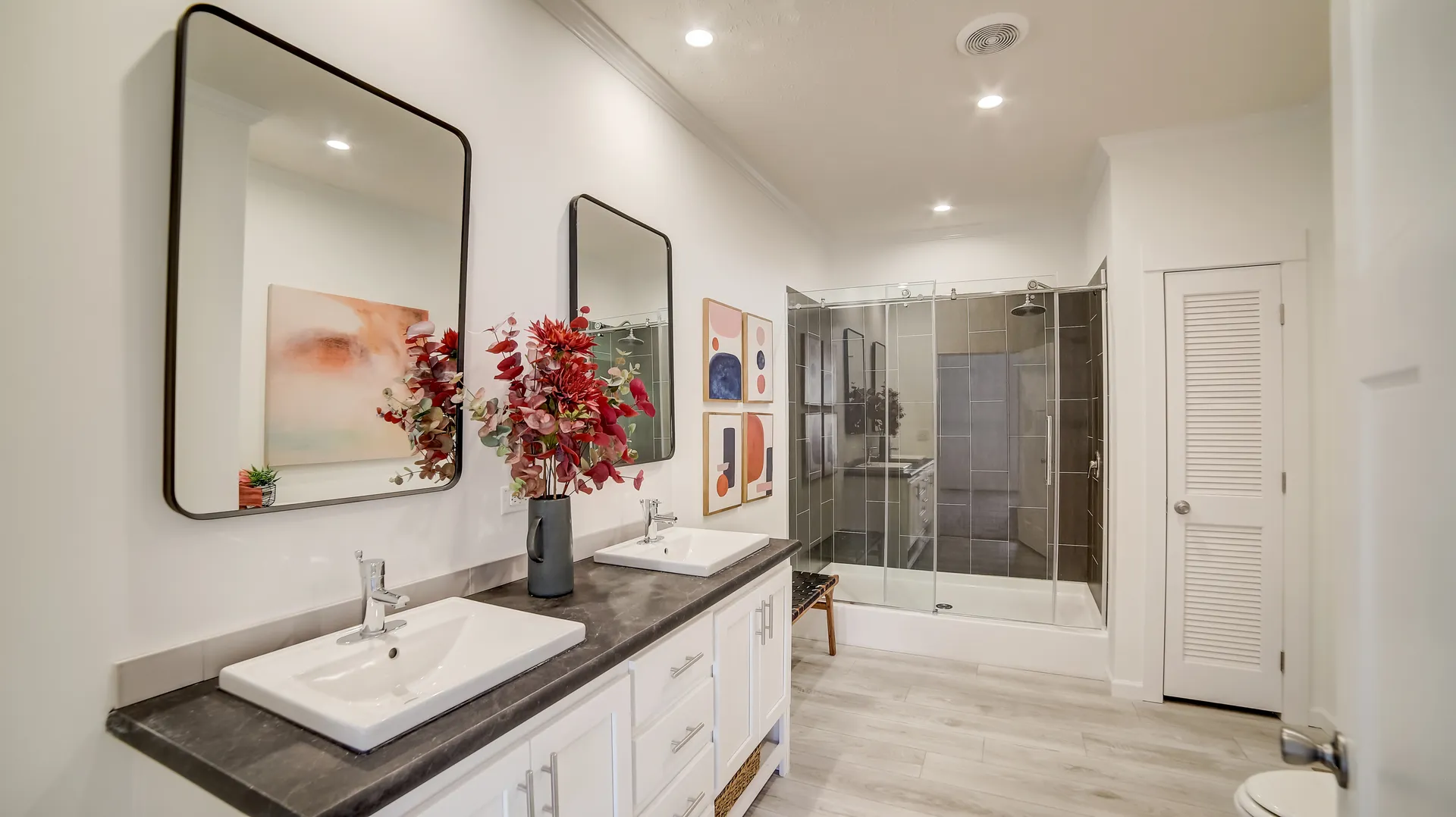 Modern bathroom with dual sinks on a black countertop and rectangular mirrors. Red foliage in a vase, abstract art, glass shower, and white walls create a sleek, fresh ambiance.