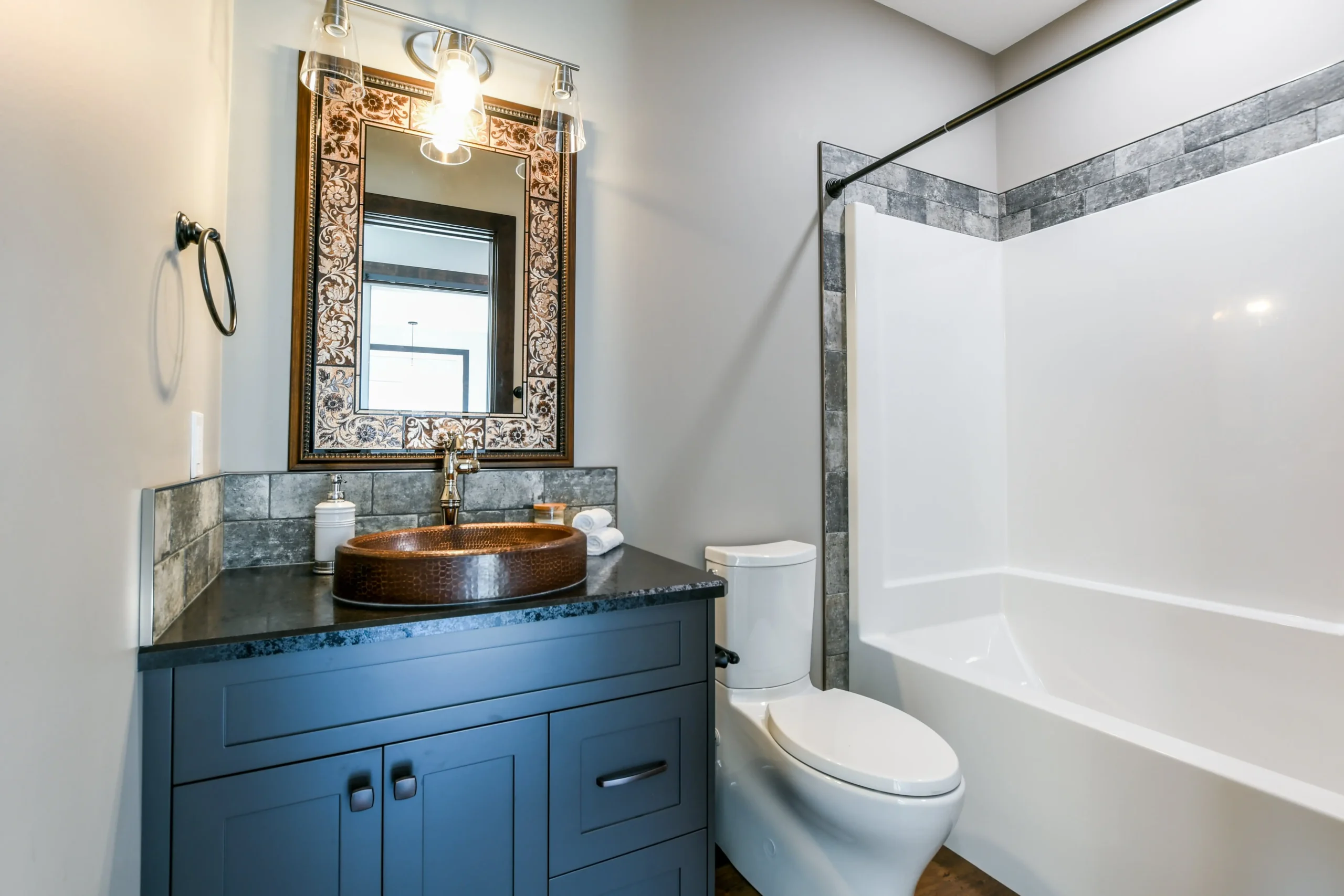 Stylish bathroom with a dark vanity, ornate mirror, vessel sink, and modern light fixture. Features a white toilet and tiled bathtub area.