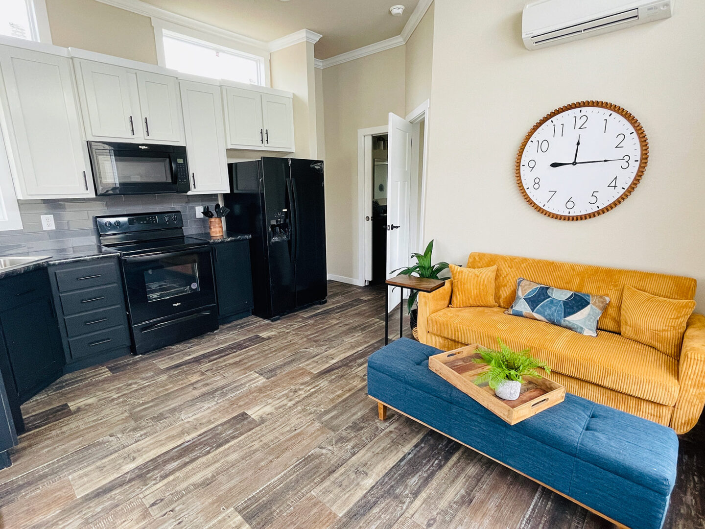 A cozy living space with a mustard yellow couch, blue ottoman, and large wall clock. Adjacent is a modern kitchen with black appliances. The ambiance is warm and inviting.